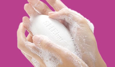 A person holds a bar of Joylux SHEbar® Intimate Soap in their hands. Their hands are sudsy and foamy.