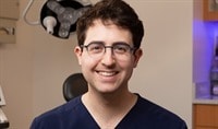 A young dermatologist with glasses, fair skin and dark brown hair wearing navy blue scrubs smiles at the camera.