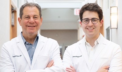Two male dermatologists, one in his 60s and one in his 30s, stand side by side in white doctor jackets smiling.