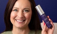 A woman with a light, neutral skin tone and dark brown hair holds a bottle of ALASTIN Skin Nectar with TriHex Plus while smiling at the camera.