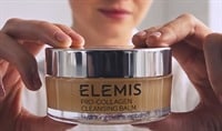 A woman smiling softly and holding a jar of ELEMIS Pro Collagen Cleansing Balm in front of her face.l