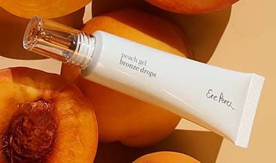 A tube of Ere Perez Peach Gel Bronzing Drops lays over peaches. Some are cut open and some are whole.
