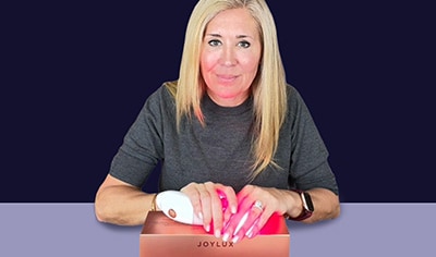 A woman with a light skin tone, brown eyes and blonde hair looks at the camera while holding a Joylux vFit device.