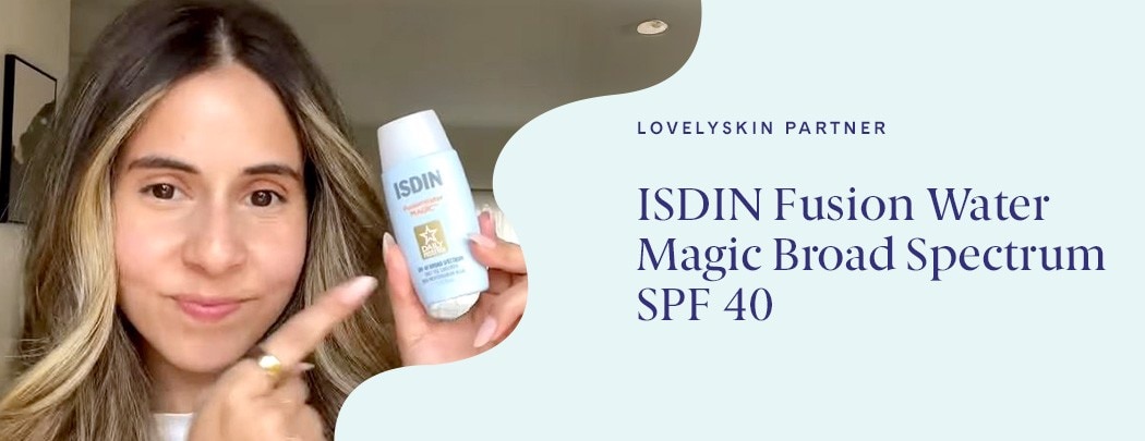 A woman with a medium skin tone and dark brown hair smiles as she points to a blue bottle of ISDIN Fusion Water Magic Broad Spectrum SPF 40 sunscreen.