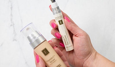 A woman with a light skin tone and pink fingernails holds the Eminence Organics Viamin C+ Serum and Vitamin C+ Eye Cream over a marble background.