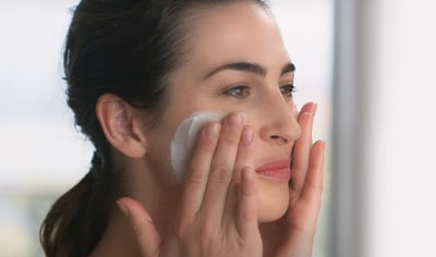 A woman with a light skin tone and dark brown hair lathers facial cleanser on her face.