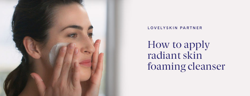 A woman with a light skin tone and dark brown hair lathers facial cleanser on her face.