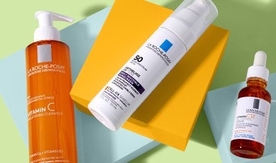 Bottles of La Roche-Posay cleanser, sunscreen and vitamin C serums laying on pastel blue and bright orange blocks on a light green background.