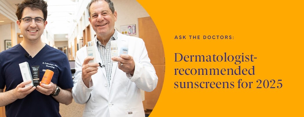 Two dermatologists, one in his 30s with dark hair &one in his 60s with light hair, stand next to one another smiling while holding various sunscreens.