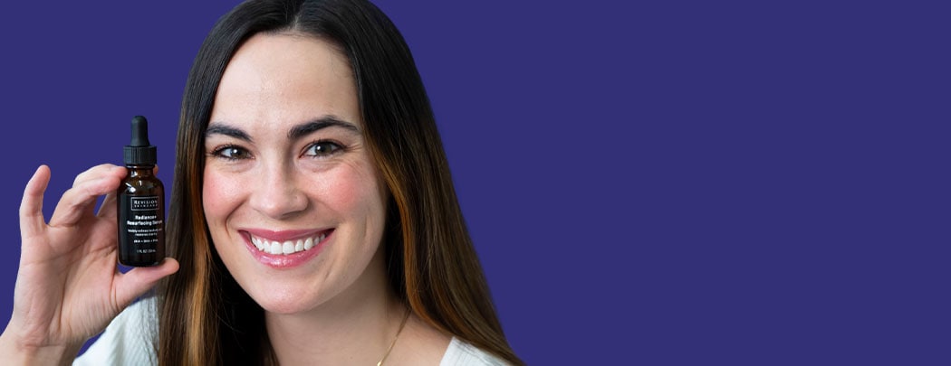 A woman with olive toned skin and dark brown hair smiles while holding a brown serum bottle that has a black label.