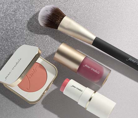 A powder blush in a compact, a cream blush in a tube, and a liquid blush in a tube lay next to a blush brush.