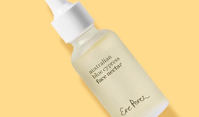 A woman with a medium skin tone and short brown hair drops Ere Perez Australian Blue Cypress Face Nectar onto her cheek.