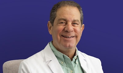 Dr. Joel SChlessinger sits in a gray chair wearing a green button up shirt in his white doctor jacket while smiling.