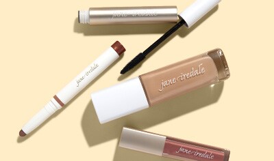 A gold mascara tube lays next to a white eyeshadow stick, a bottle of liquid foundation and a rose-colored lip gloss.