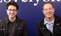 Two dermatologists, one in his 30s with dark hair & one in his 60s with light hair, sit in chairs as they smile at the camera.