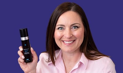 A woman with fair skin and dark hair smiles while holding a dark bottle of SkinCeuticals Advanced RGN-6. She wears a pink shirt.