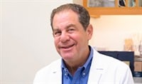 Dr. Joel Schlessinger, a board-certified dermatologist, sits in a patient room in a white jacket as he smiles.
