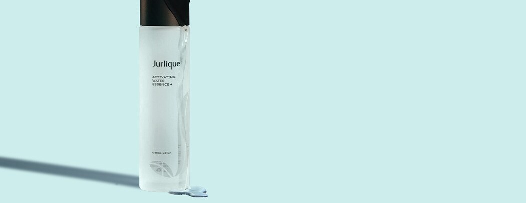 A clear bottle of Jurlique Activating Water Essence+ with liquid running over the bottle and black cap on a soft blue background.