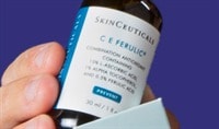 A person with an olive skin tone holds a bottle of SkinCeuticals C E Ferulic, almost as if they're pulling it out of the box.
