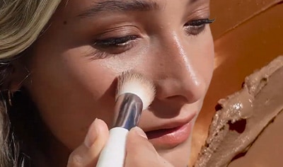 A woman with a light skin tone and blonde hair presses a brush to her cheek. There are swatches of the Ere Perez Cacao Bronzing Pot behind her.