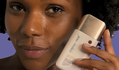 A woman with a deep skin tone and dark brown hair smiles while holding a bottle of Jane Iredale Glow Fixation.