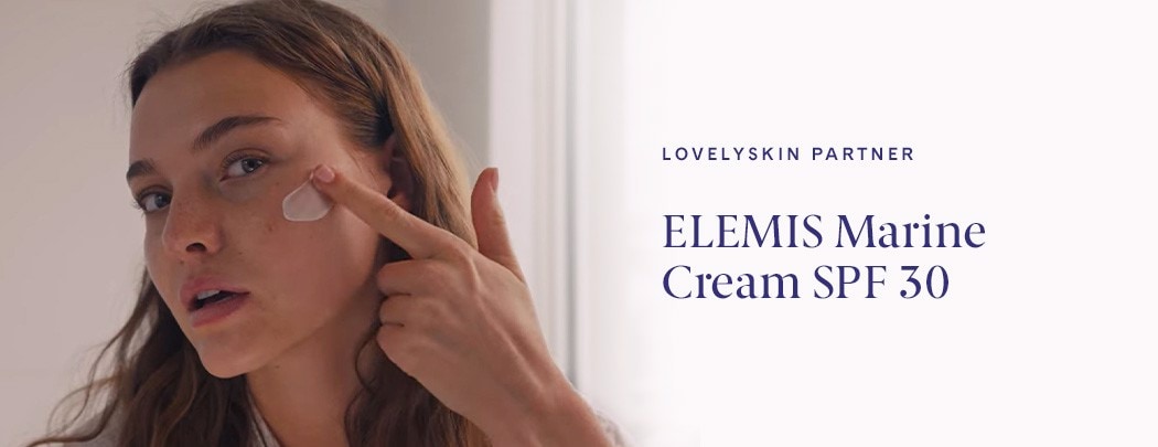 A woman with curly red hair applying a swatch of white ELEMIS Marine Cream SPF 30 across her cheek with her fingertip.