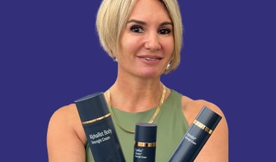 A woman with a light skin tone and a short blonde bob holds three skinbetter science AlphaRet products while smiling.
