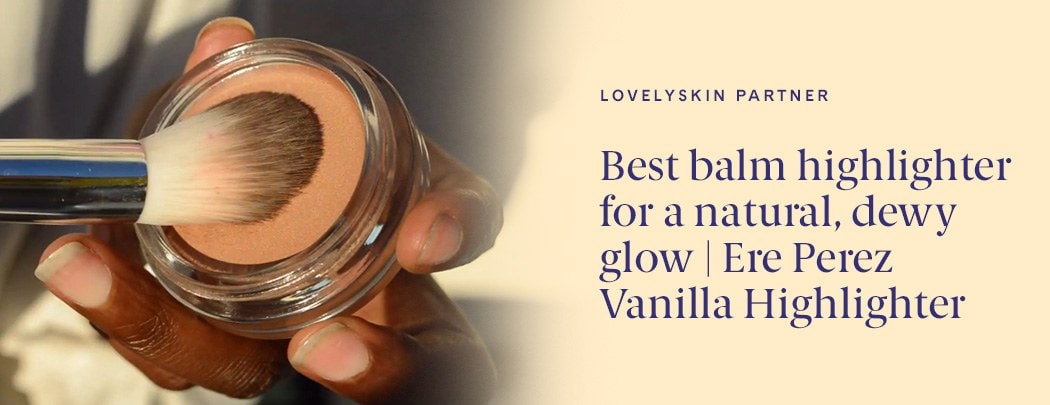 A person with a deep skin tone holds a container of Ere Perez Vanilla Highlighter in her hand while pressing a brush into it.