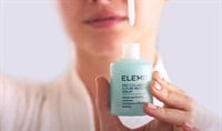 A white woman holding a bottle of ELEMIS Pro Collagen Future Restore Serum with a dropper suspended above it.