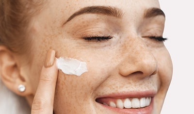 A fair-skinned model with freckles with her eyes closed applying moisturizer to her cheek.