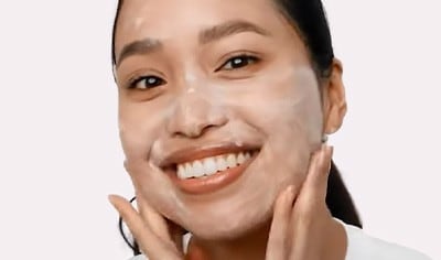 Woman of East Asian descent smiling at camera while blending foaming cleanser into face