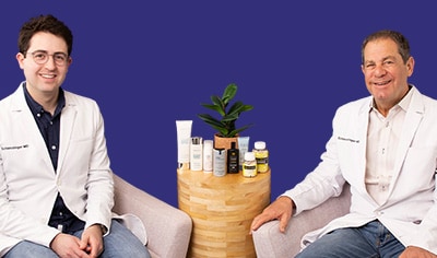Drs. Joel and Daniel Schlessinger sit next to one another. Between them is a wood table with an array of sunscreens. They are both smiling.