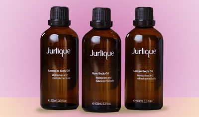 Three Jurlique Body Oils in natural brown glass sit next to one another on a yellow table with a pink background.