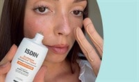 A woman with a light skin tone and brown hair holds a bottle of ISDIN Eryfotona Actinica Tinted Mineral SPF 50 while showing a swatch on her fingers.