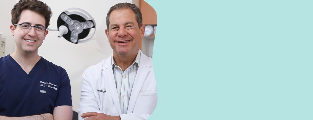 Two dermatologist, one in his 30s with fair skin & dark hair & one in his 60s with light brown hair smile while crossing their arms.