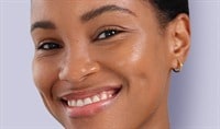 A woman with a deep skin tone smiles at the camera. Her skin looks dewy and radiant.