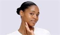 A woman with a deep skin tone and dark hair turns her face to the side while gently touching her jaw and smiles softly.