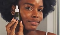 A black woman smiling softly and holding a bottle of ELEMIS Superfood Midnight Facial Oil next to her face.