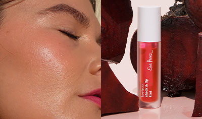 A woman with medium skin tone and brown hair shows her lips and cheeks with a pink hue. A tube of Beetroot Cheek & Lip Tint is pictured next to her.