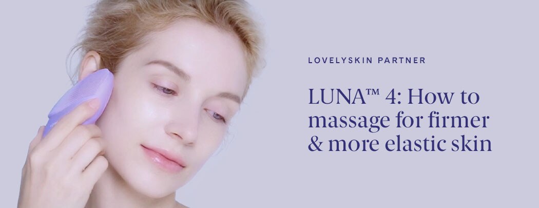 A woman with fair skin and blonde hair holds a lavender skin care device against her cheek while looking downward.