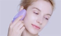 A woman with fair skin and blonde hair holds a lavender skin care device against her cheek while looking downward.