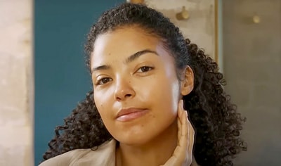 A woman with a medium to deep skin tone and curly brown hair looks into the camera, as if into a mirror, with her hand gently placed below her jaw.