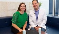 A man in a doctor's coat and a woman in a green shirt sitting side by side and smiling at the camera.