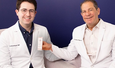 Drs. Joel and Daniel Schlessinger sit next to one another while smiling. Dr. Joel holds a box of EltaMD UV Clear Blemish-Prone & Oil Balancing SPF 50.