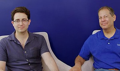 A man in his 30s with fair skin & dark brown hair sits in a chair next to a man in his 60s with a medium skin tone & light brown hair.