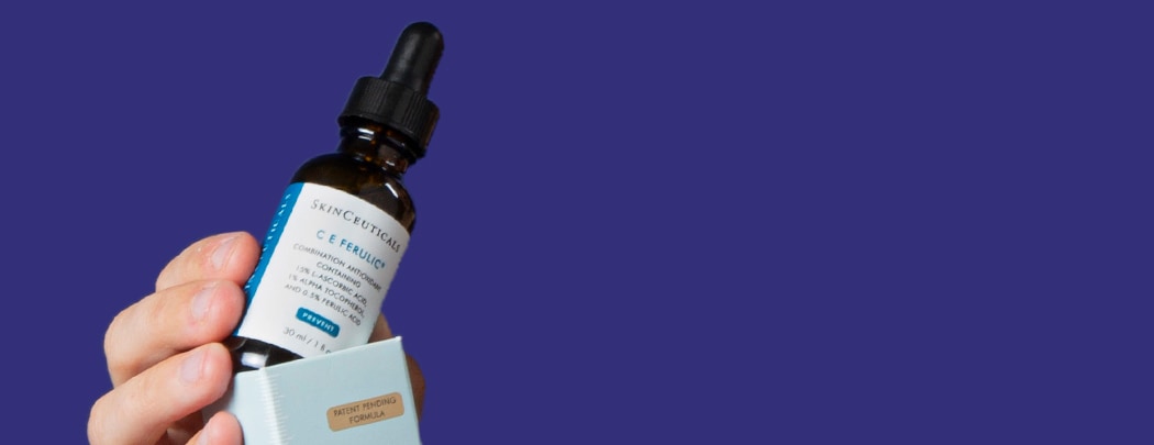 A person with an olive skin tone holds a bottle of SkinCeuticals C E Ferulic, almost as if they're pulling it out of the box.