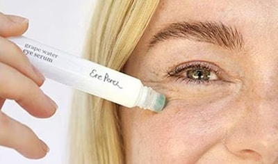 A woman with fair skin and blonde hair presses the Ere Perez Grape Water Eye Serum to her skin while smiling.