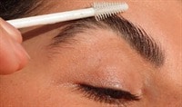 A person with medium, olive-toned skin holds a Ere Perez Aloe Gel Lash & Brow Mascara brush to their eyebrow.