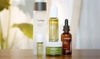 Four products in glass bottles are arranged aesthetically on a table with plants in the background.
