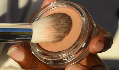 A person with a deep skin tone holds a container of Ere Perez Vanilla Highlighter in her hand while pressing a brush into it.